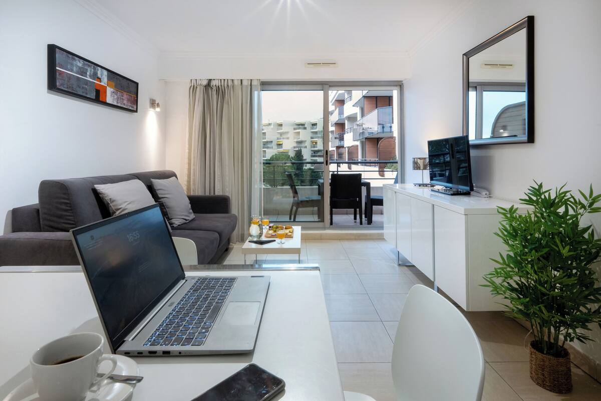 A modern living room with a laptop on a white table, a grey sofa, a white coffee table with drinks, and a balcony with a view of buildings.