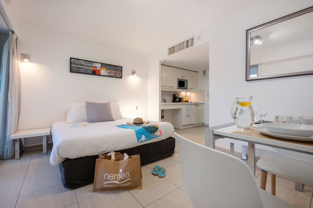 A modern hotel room with a bed, bedside table, and a view into the kitchen area.