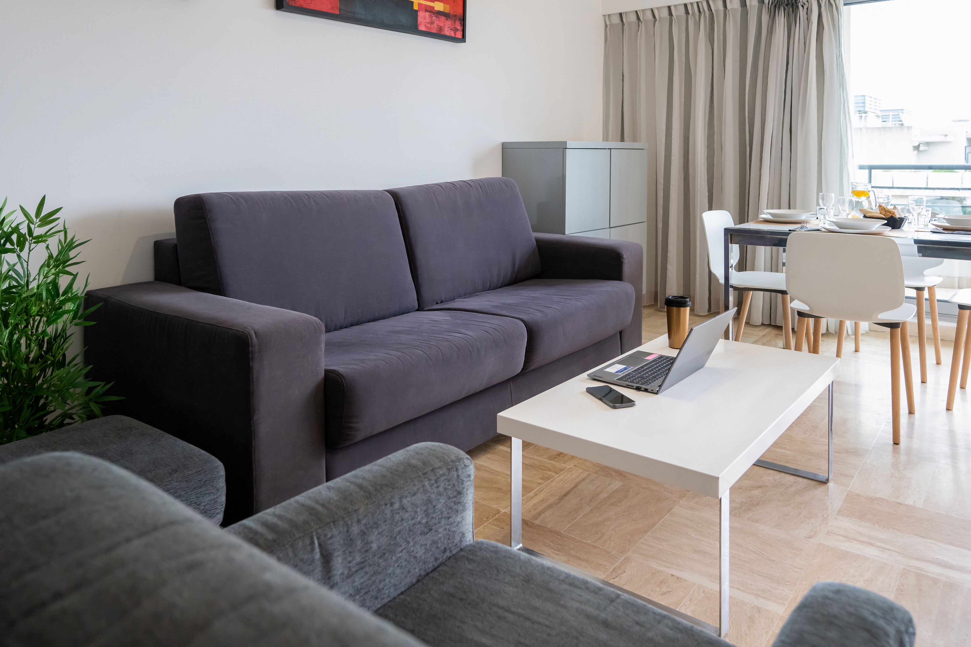 A modern living room with a grey sofa, a white coffee table with a laptop, and a dining area with a white table and chairs.