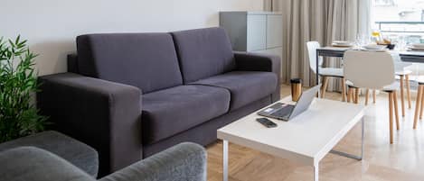 A modern living room with a grey sofa, a white coffee table with a laptop, and a dining area with a white table and chairs.