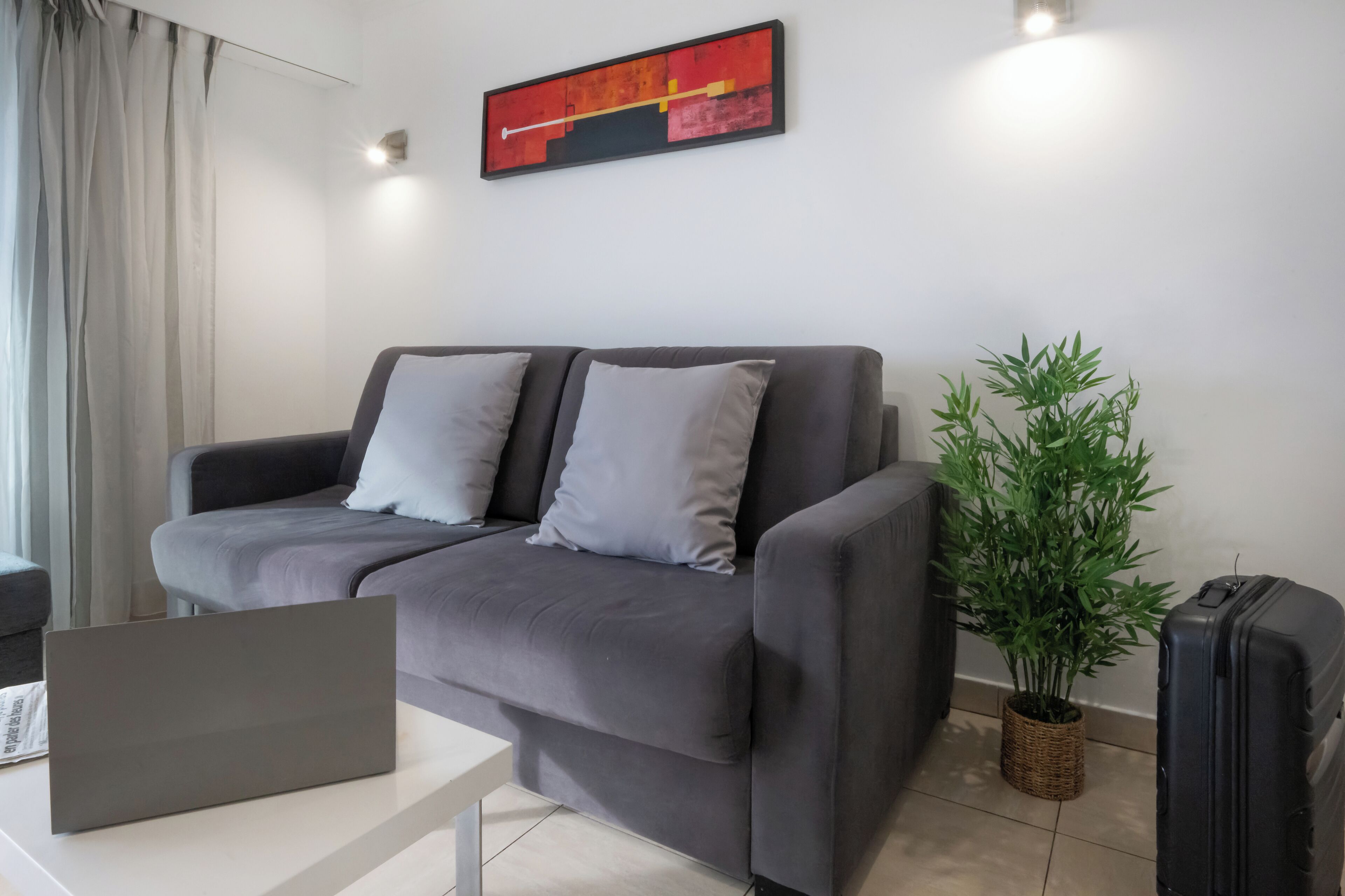 A modern living room with a grey sofa, a laptop on a white table, a potted plant, and a suitcase.