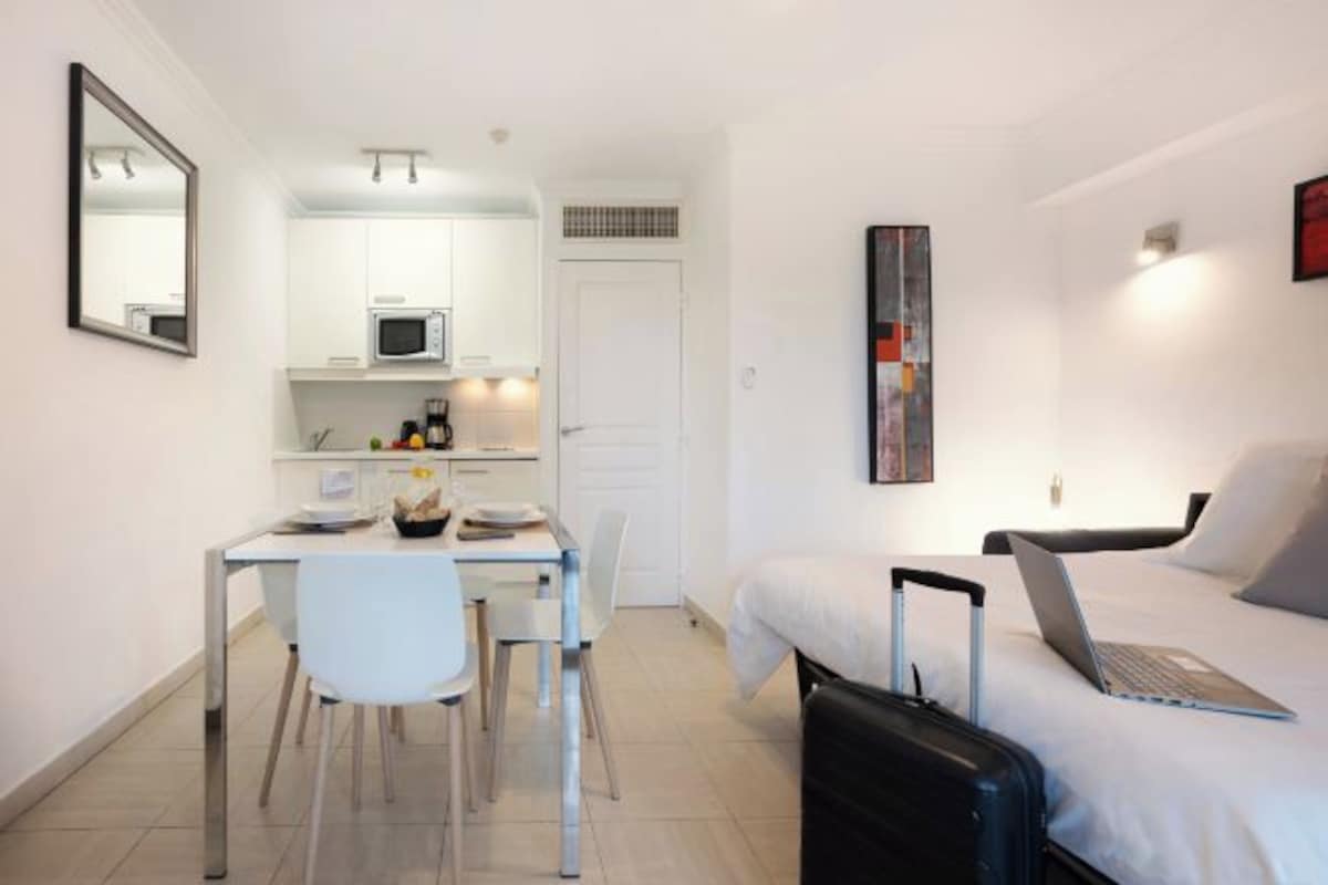 A compact hotel room with a bed, a desk, a chair, a microwave, and a suitcase.