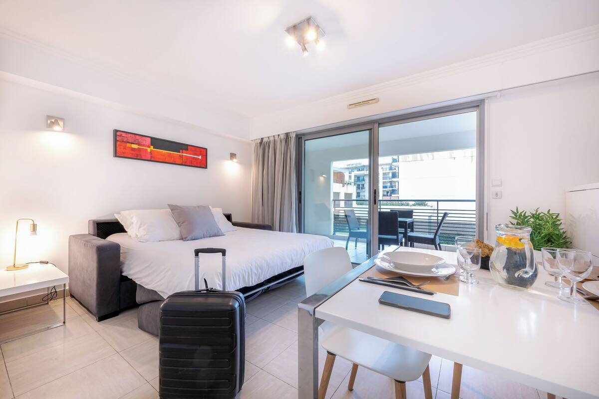 A modern hotel room with a bed, a desk, a chair, a suitcase, and a balcony with a view.