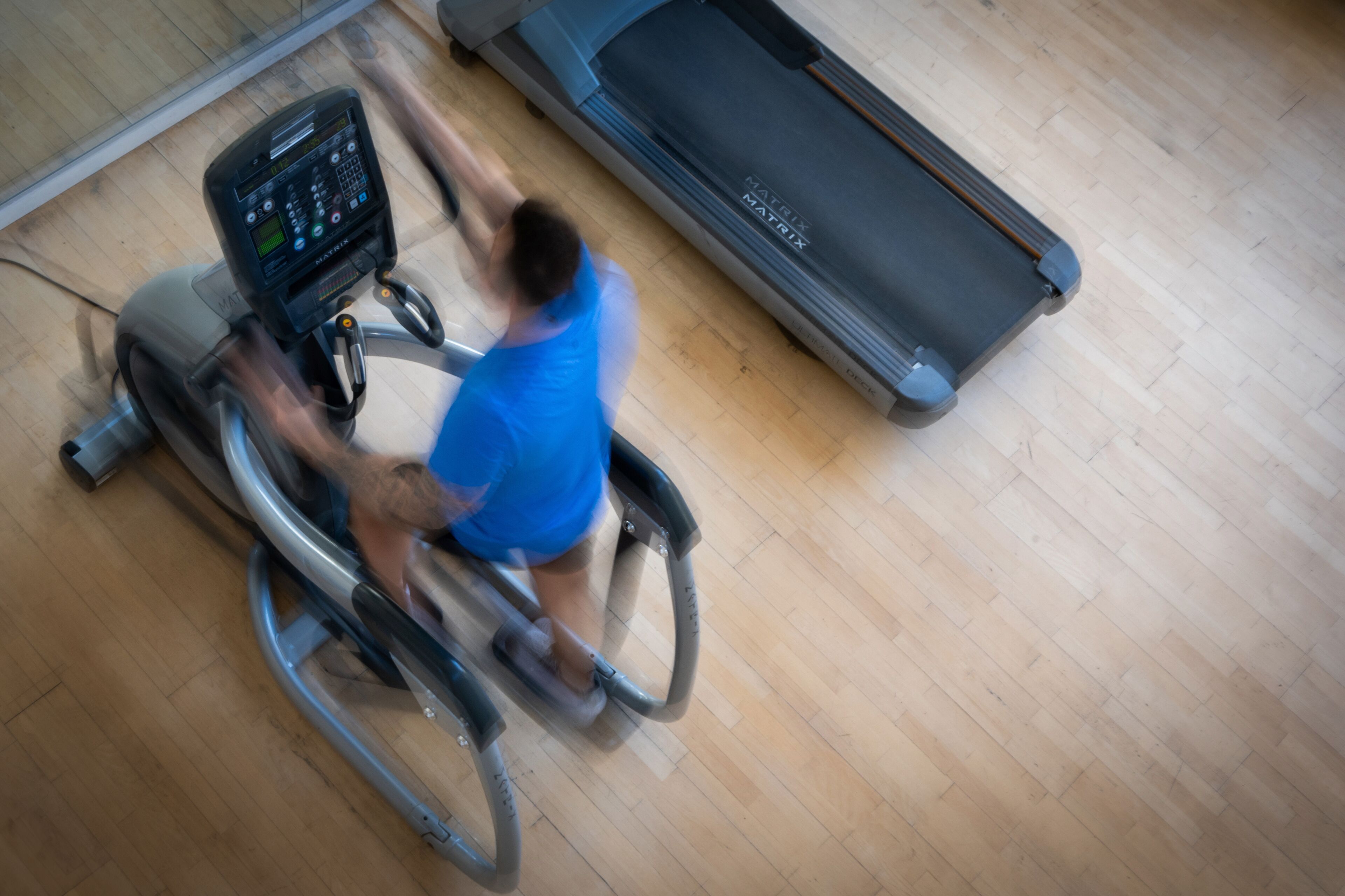 Une personne fait de l’exercice sur un vélo d’appartement dans une salle de sport, avec un tapis de course en arrière-plan.