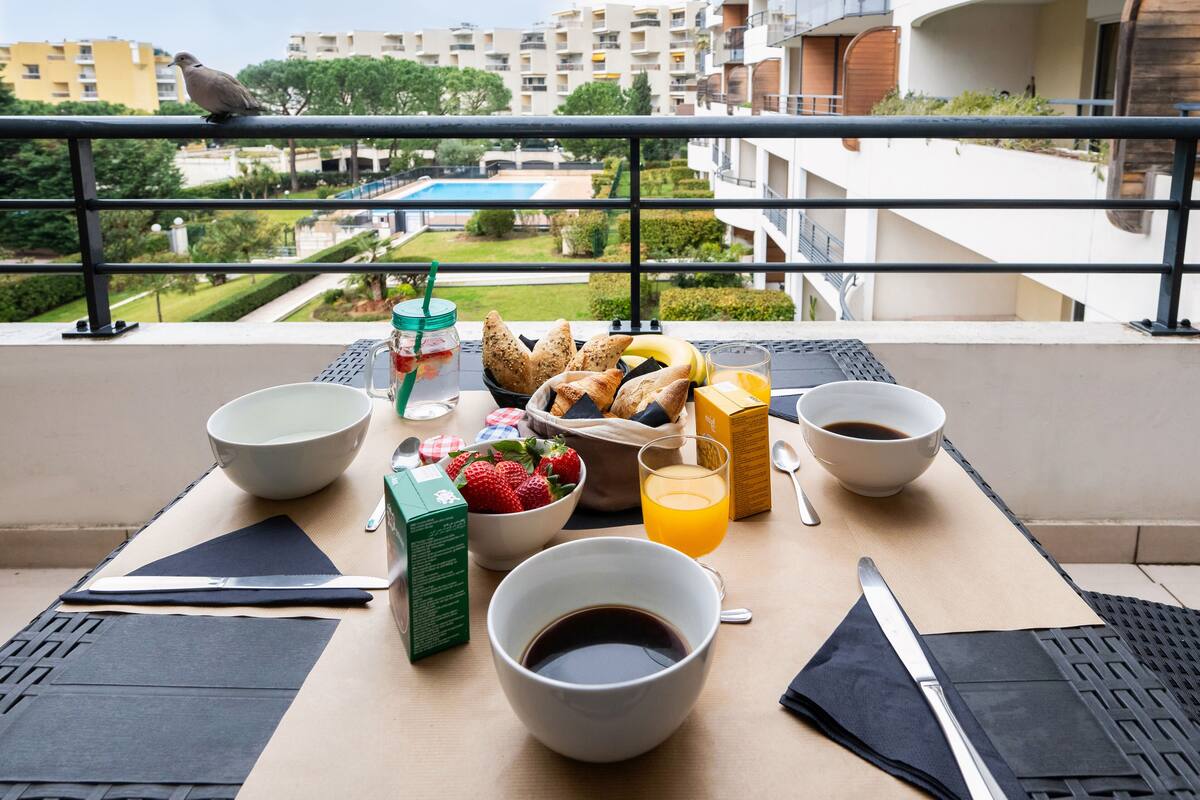 A balcony table with breakfast items, a view of a swimming pool, and apartment buildings in the background.