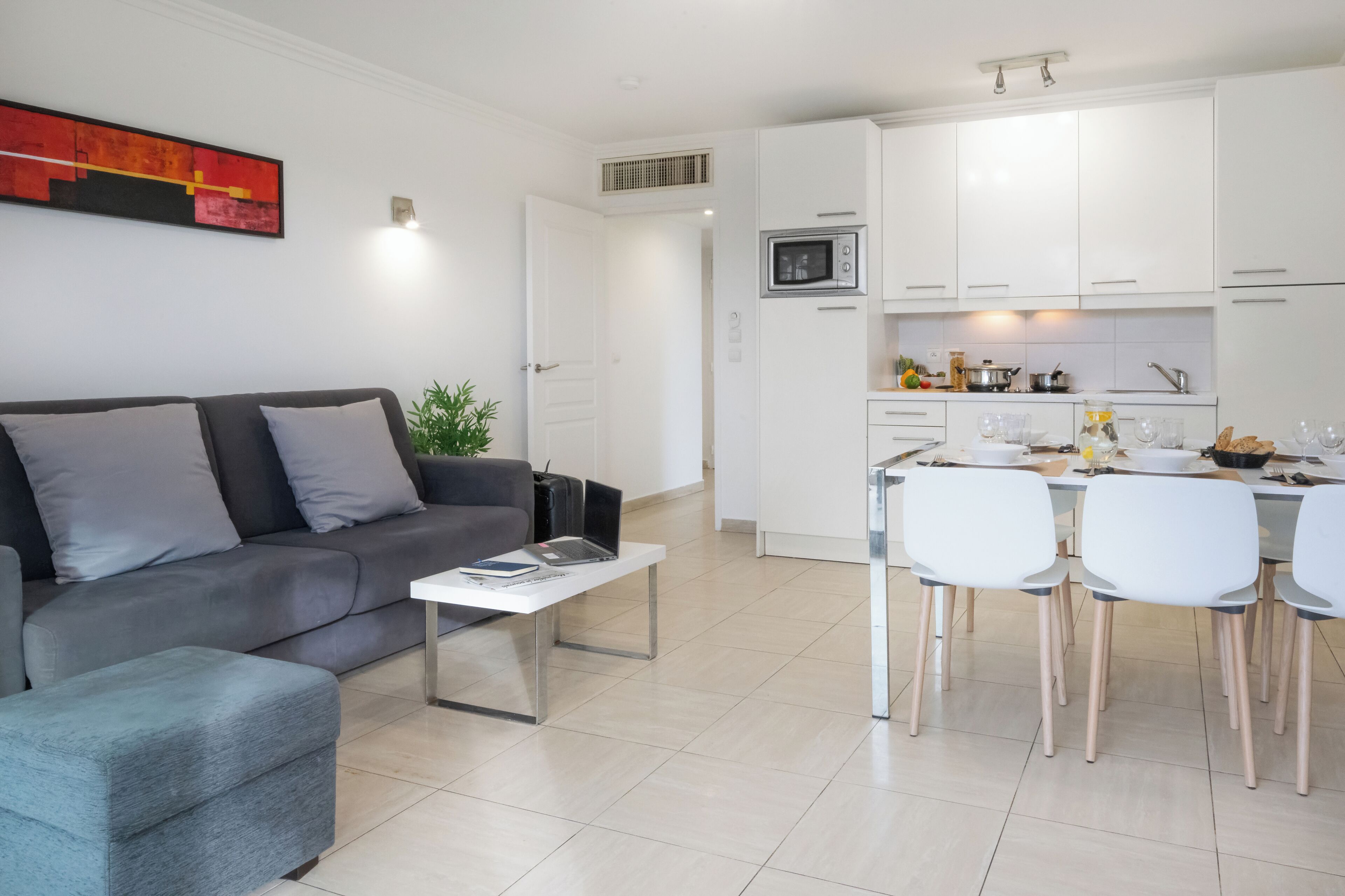 A modern living room with a grey sofa, a coffee table, and a dining area with white chairs and a kitchen in the background.