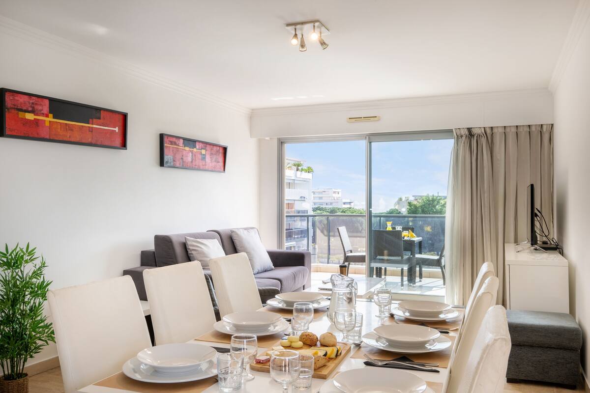 A dining area with a table set for four, a grey sofa, and a balcony with a view of buildings and greenery.