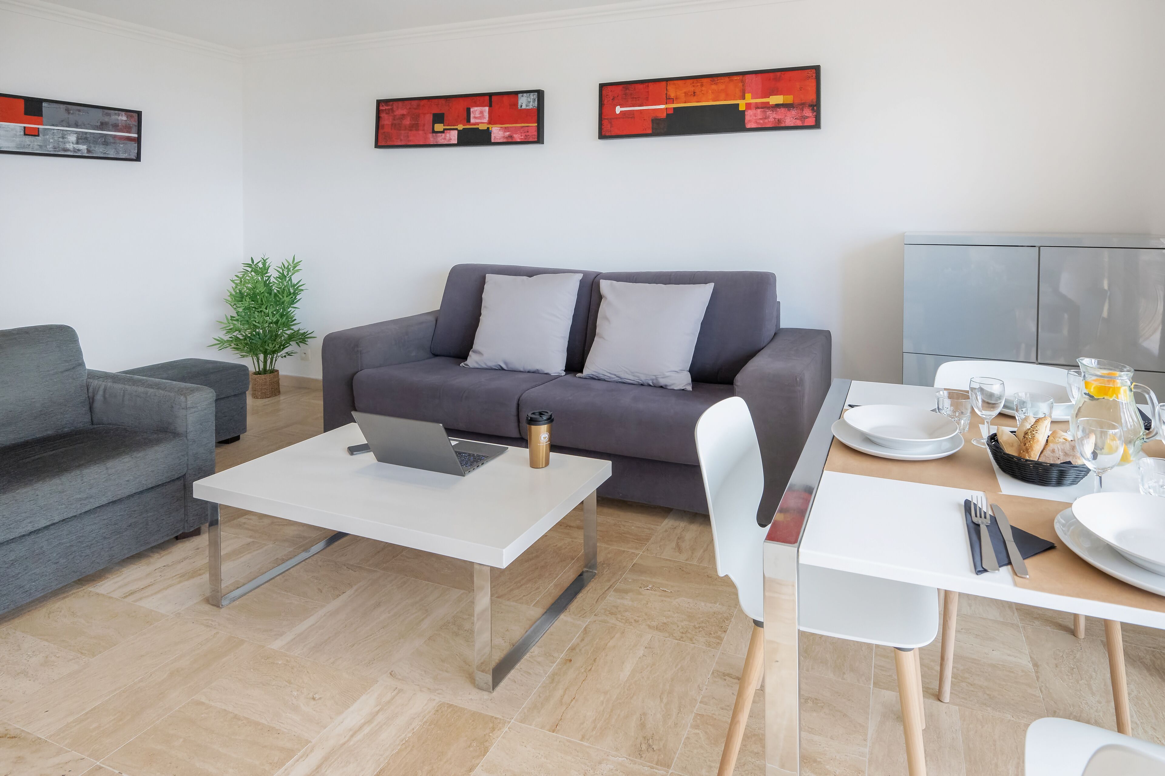 Una sala de estar moderna con un sofá gris, una mesa de centro blanca con una laptop, y un área de comedor con una mesa blanca dispuesta para dos.