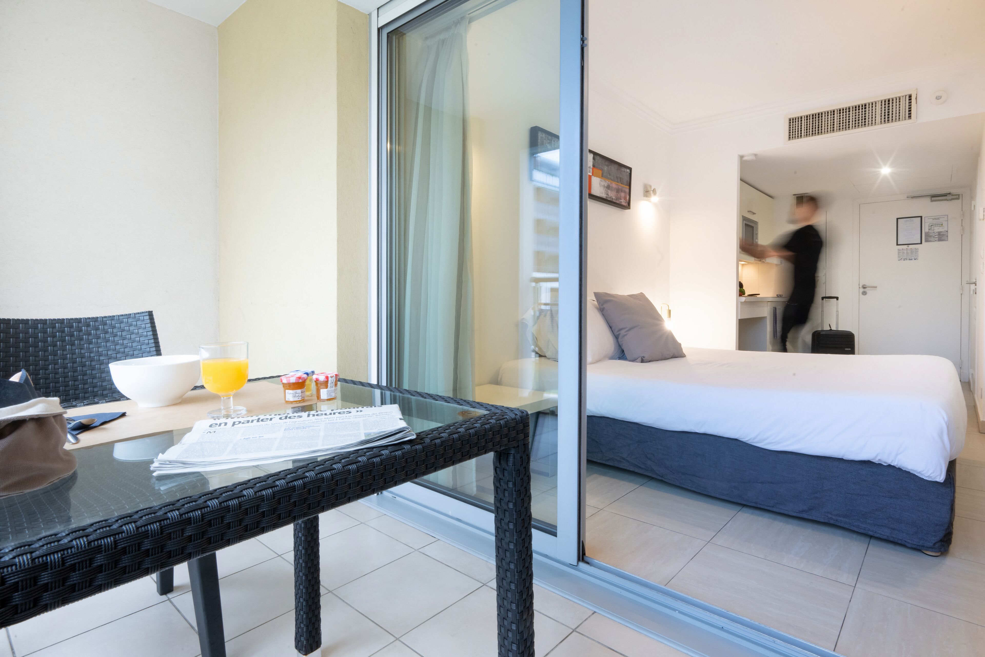 A hotel room with a glass door leading to a balcony, a bed with white bedding, a small table with a newspaper, and a bowl of orange juice.