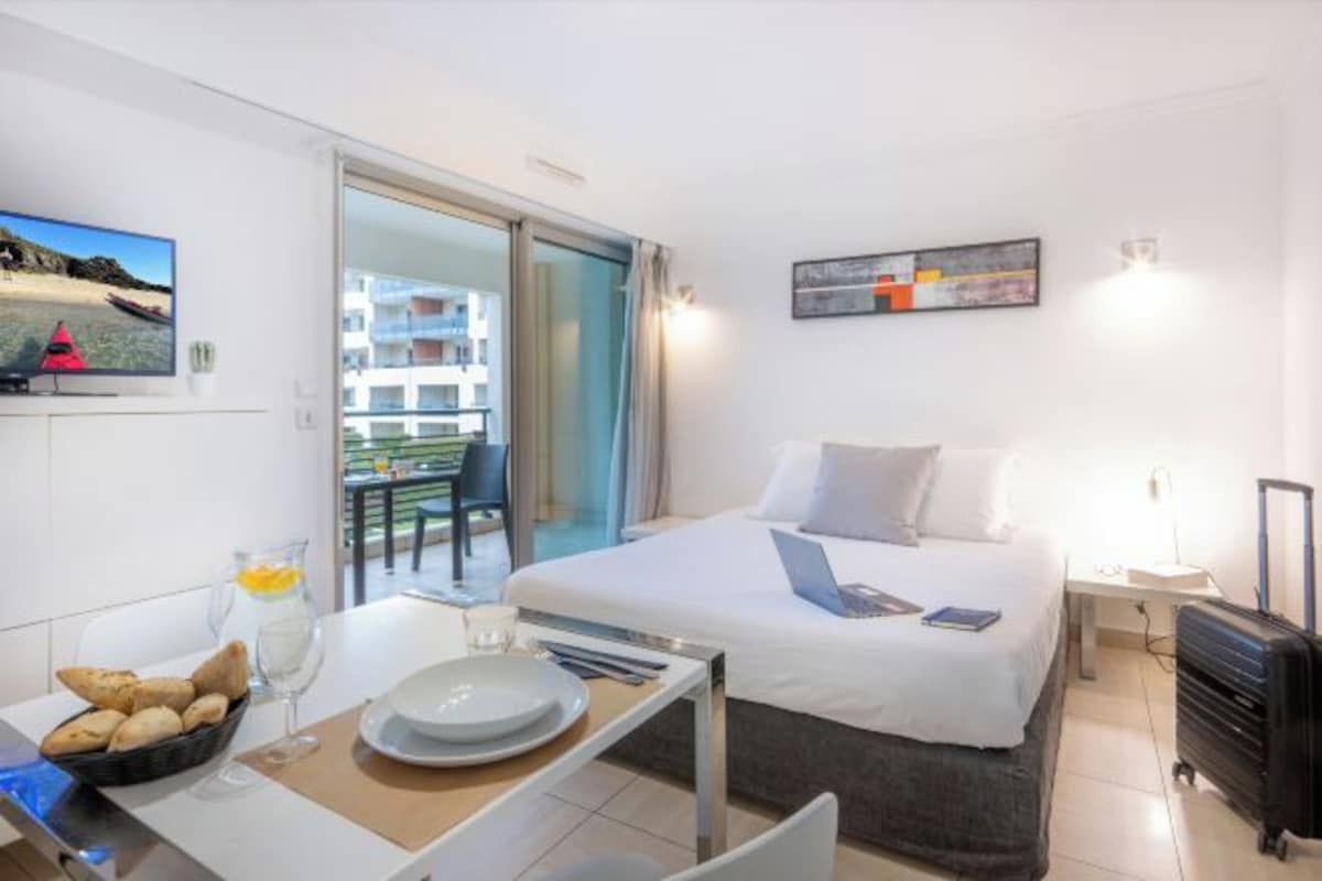 A hotel room with a bed, a desk with breakfast items, a laptop, a TV, and a balcony with a view.