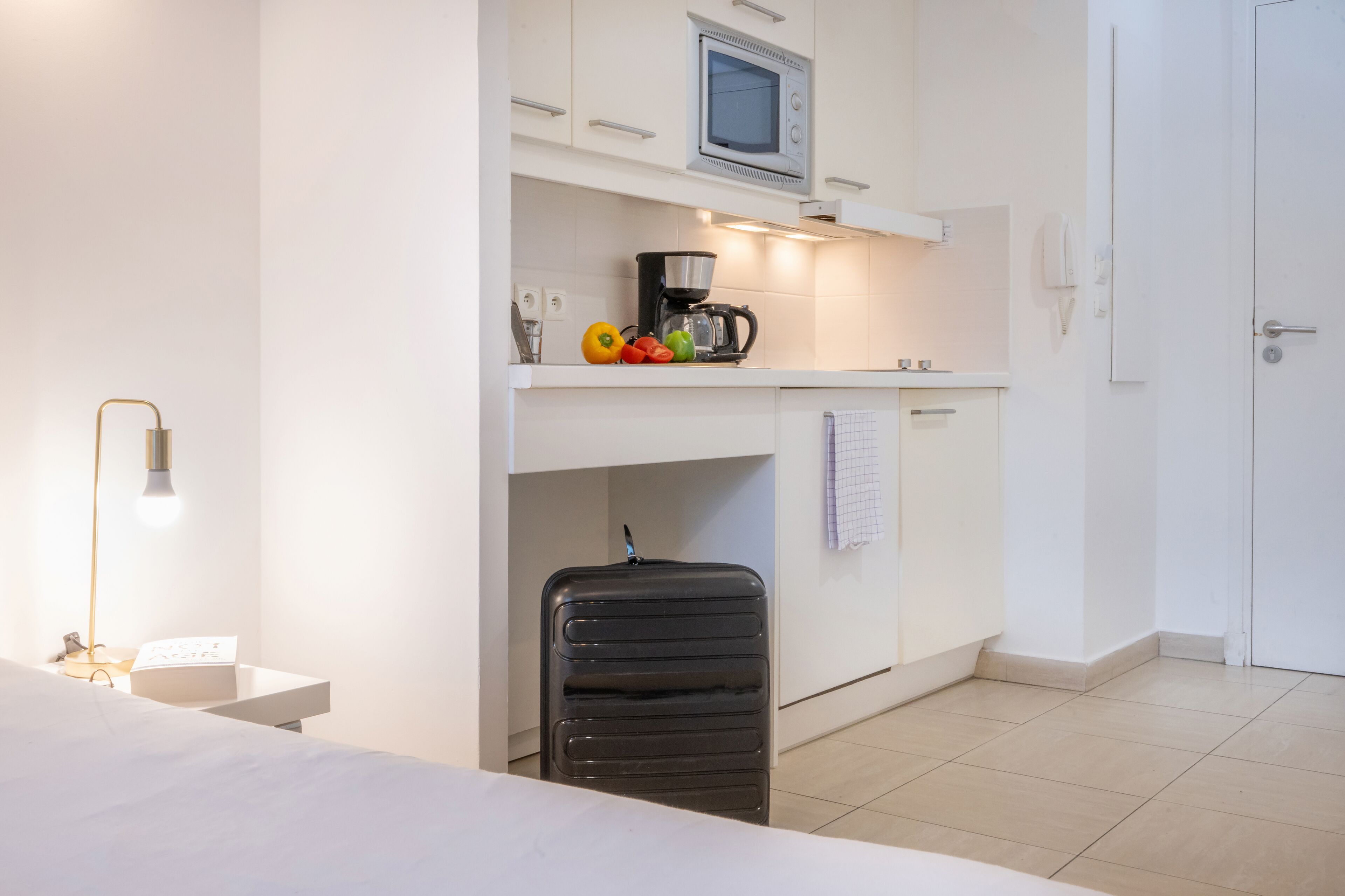 Une chambre d’hôtel de taille réduite, équipée d’une kitchenette, d’un lit, d’une lampe de chevet et d’un espace pour une valise.