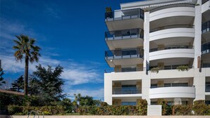 A swimming pool with a view of a multi-story building with balconies and a palm tree in the background. - Nemea Appart Hotel Le Lido Cagnes sur Mer (Cagnes-sur-Mer)