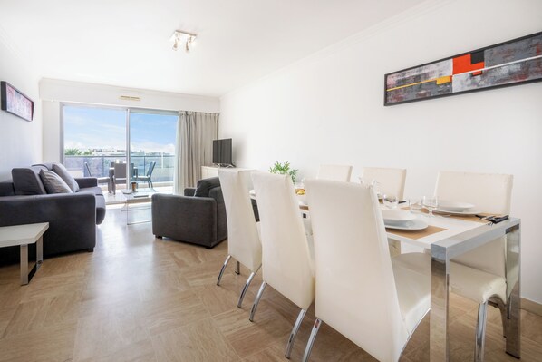 A modern living room with a sofa, a dining table set for four, and a balcony with a view. - Nemea Appart Hotel Le Lido Cagnes sur Mer (Cagnes-sur-Mer)