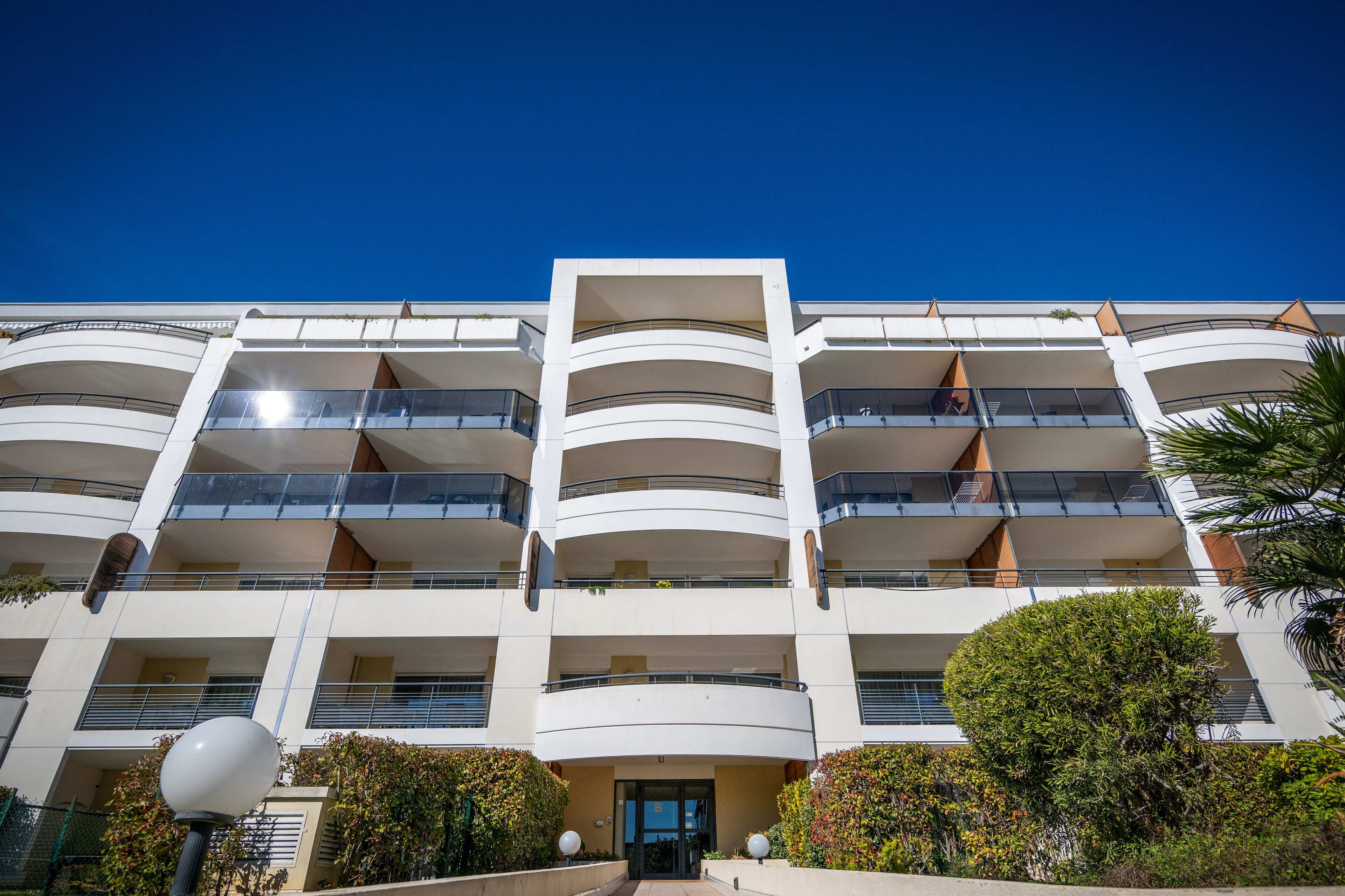 A multi-story building with balconies, a central entrance, and surrounding greenery.