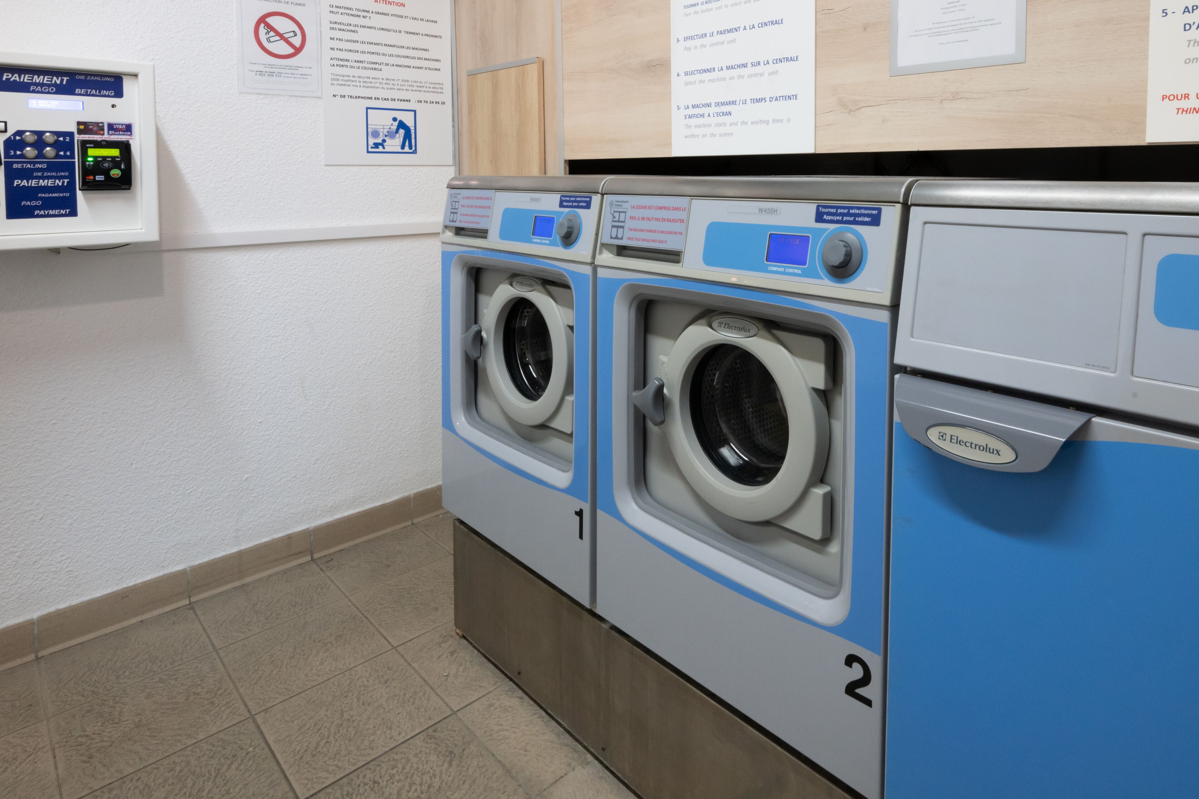 A laundry room with two front-loading washing machines, a payment terminal, and informational posters on the wall.