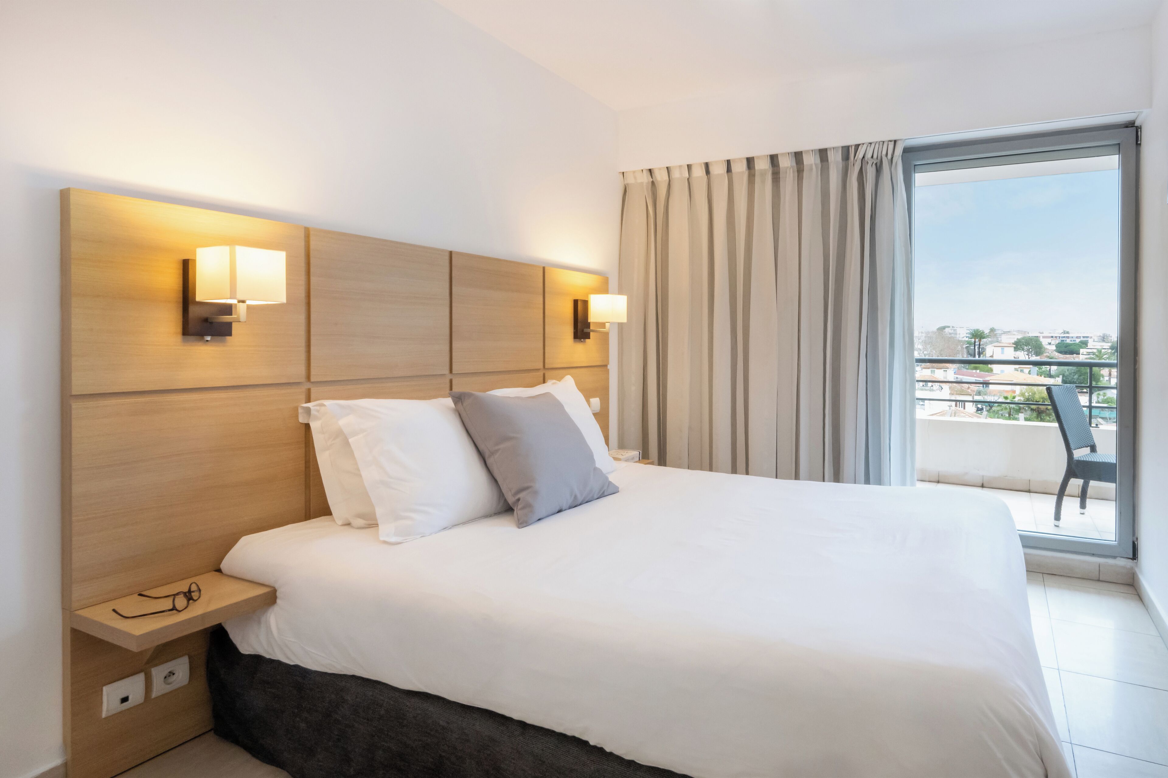 Habitación de hotel con una cama grande, dos lámparas de noche, un pequeño estante con vasos y vistas a la ciudad a través de un ventanal amplio.