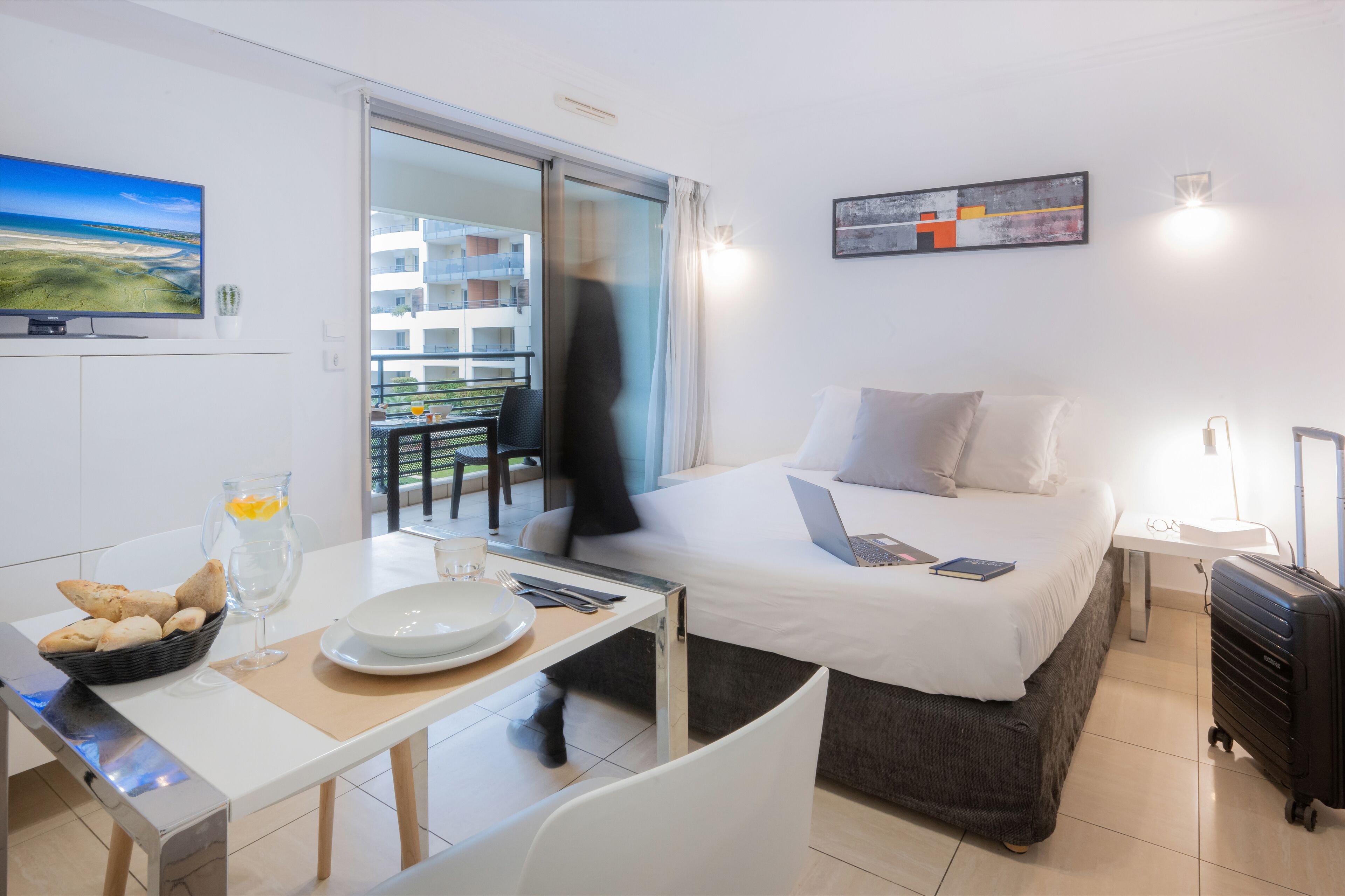 A modern hotel room with a bed, a desk with breakfast items, a laptop, and a view of the balcony and building.