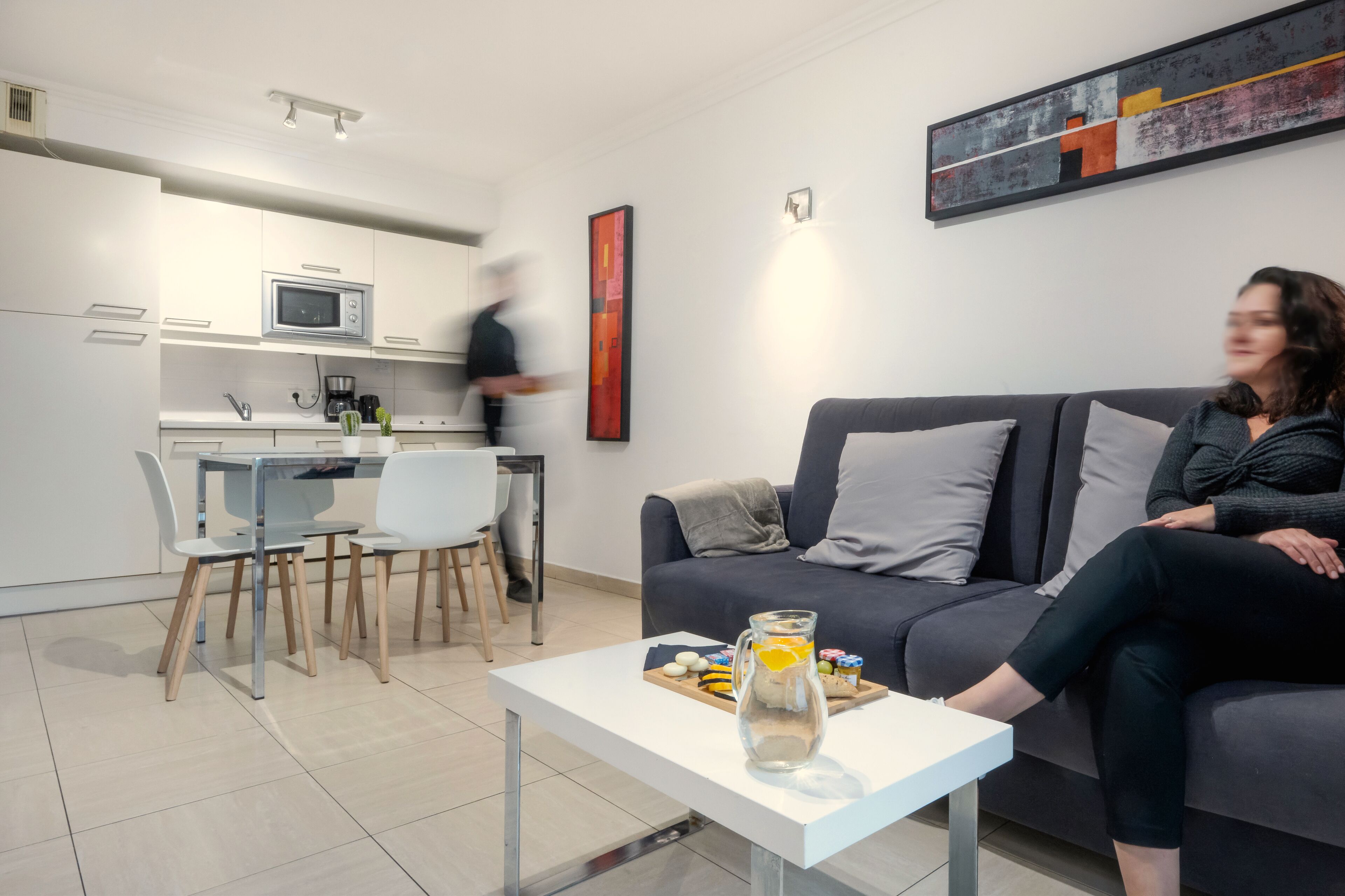 A modern living room with a grey sofa, a coffee table with a pitcher and snacks, and a kitchen area with white cabinets and a microwave.