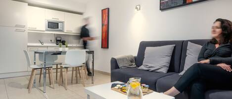 A modern living room with a grey sofa, a coffee table with a pitcher and snacks, and a kitchen area with white cabinets and a microwave.