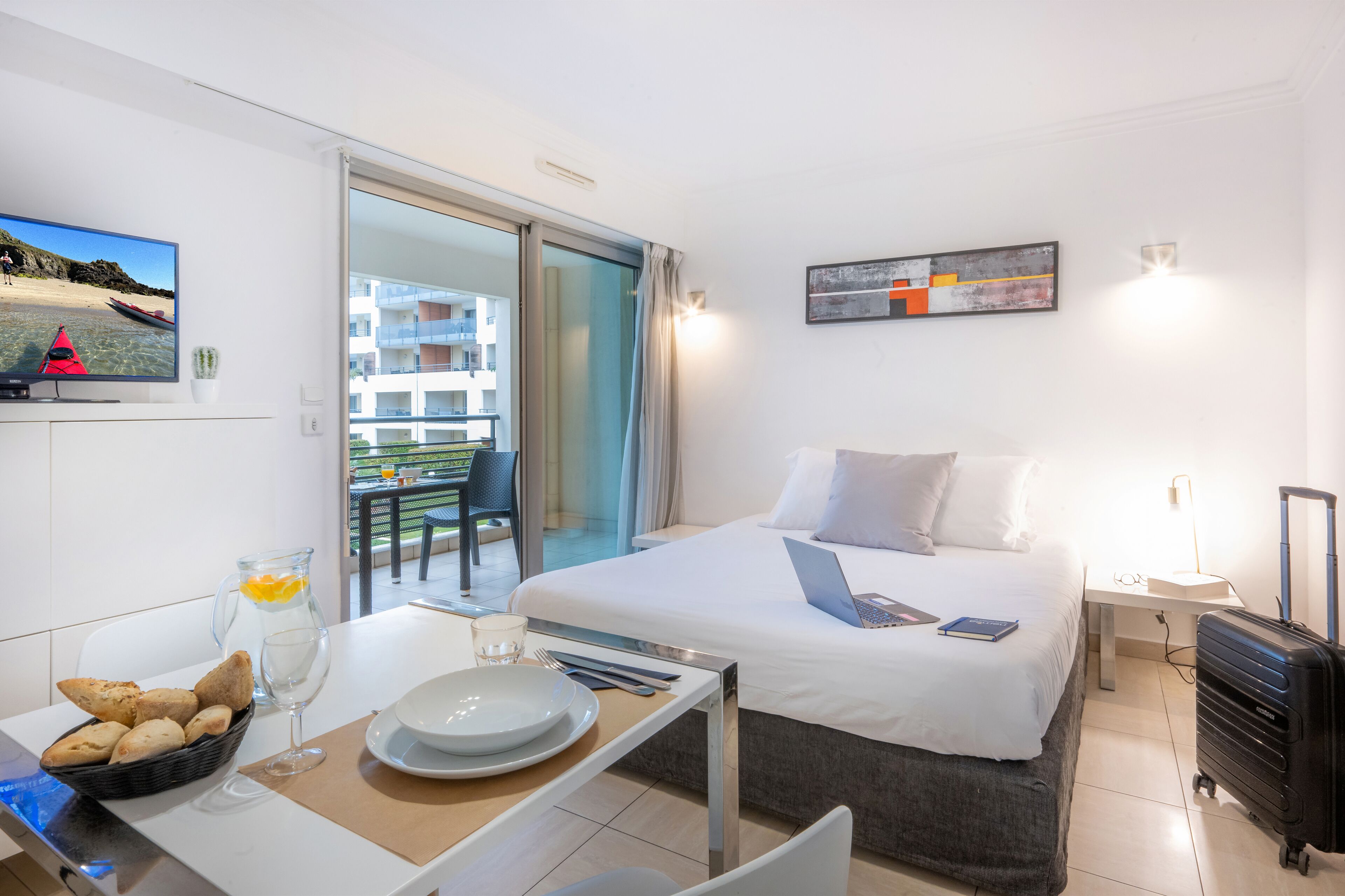 A hotel room with a bed, a desk with breakfast items, a laptop, and a suitcase.