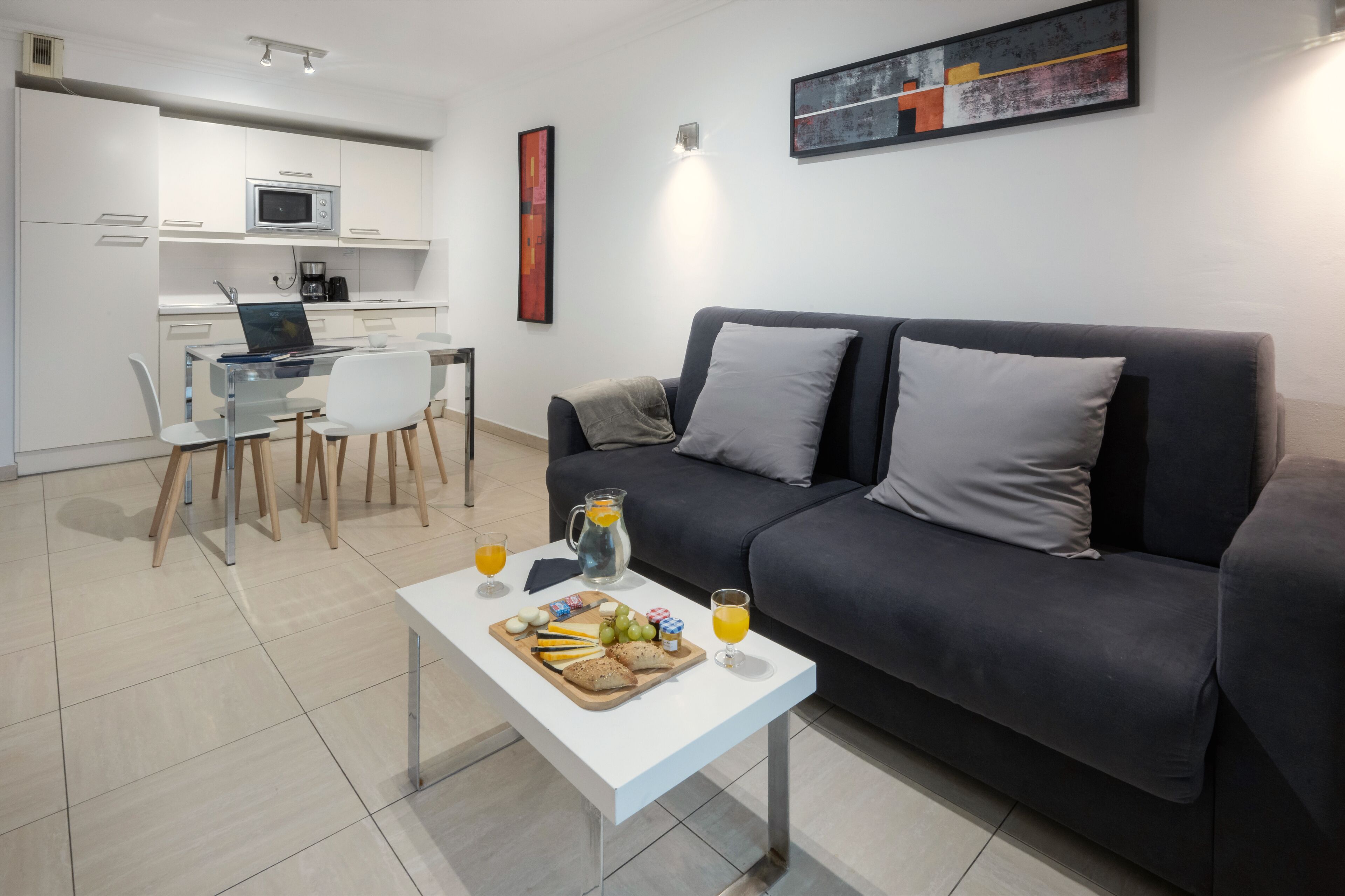 A modern living room with a dark grey sofa, a white coffee table with breakfast items, a kitchen area with white cabinets and a microwave, and a framed abstract painting on the wall.