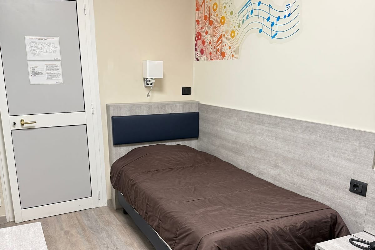 A single bed with a headboard, a bedside table, a wall-mounted light, and a door with a notice.