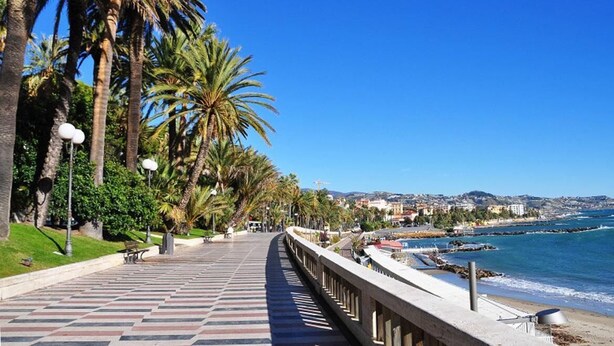 A seaside promenade with palm trees, a tiled walkway, and a view of the ocean and distant buildings. - Hotel Esperia (Sanremo)