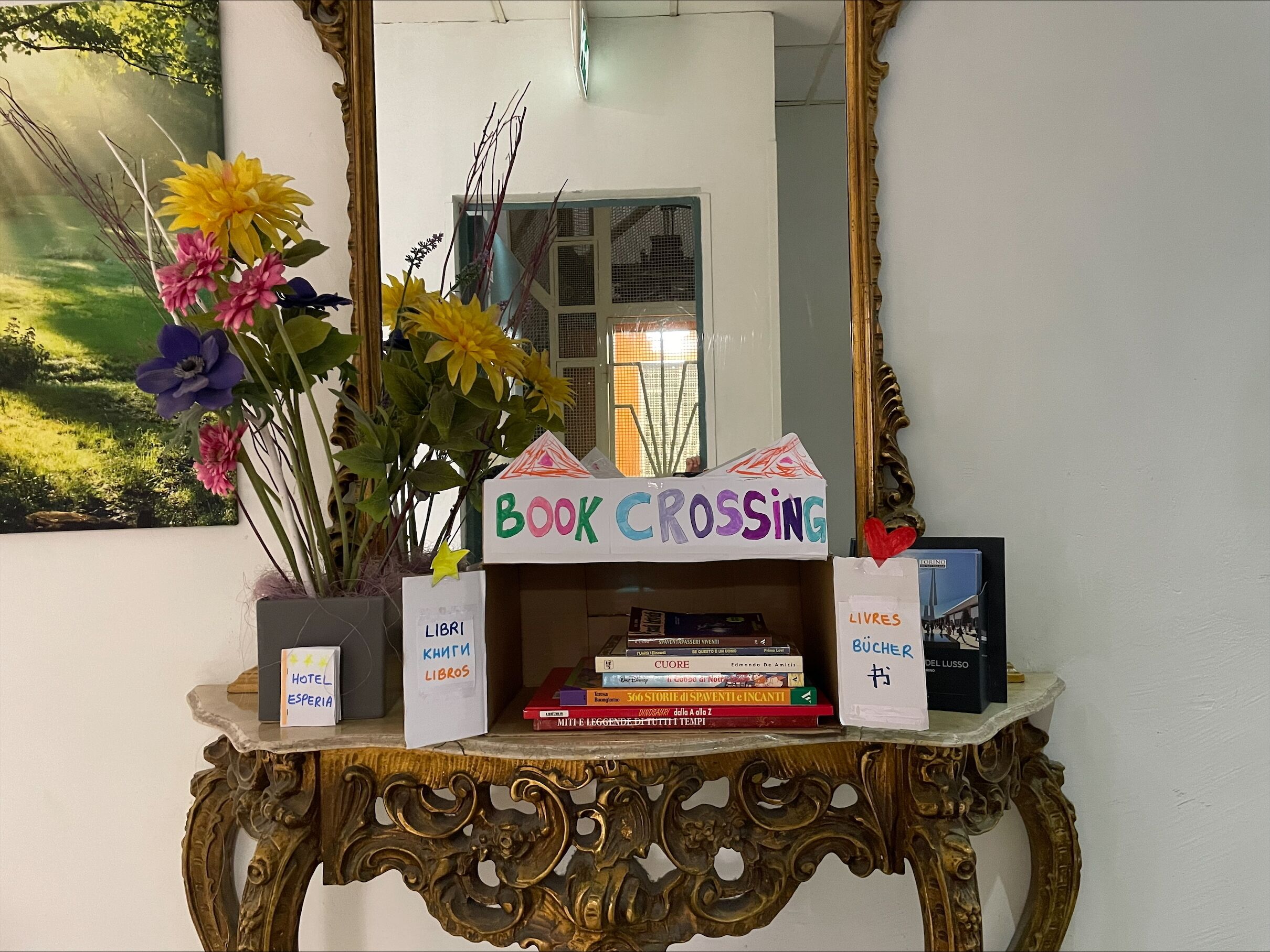 Un mostrador de recepción de hotel con un espejo, una planta en maceta con flores, un letrero que dice "BOOK CROSSING" y una pila de libros.