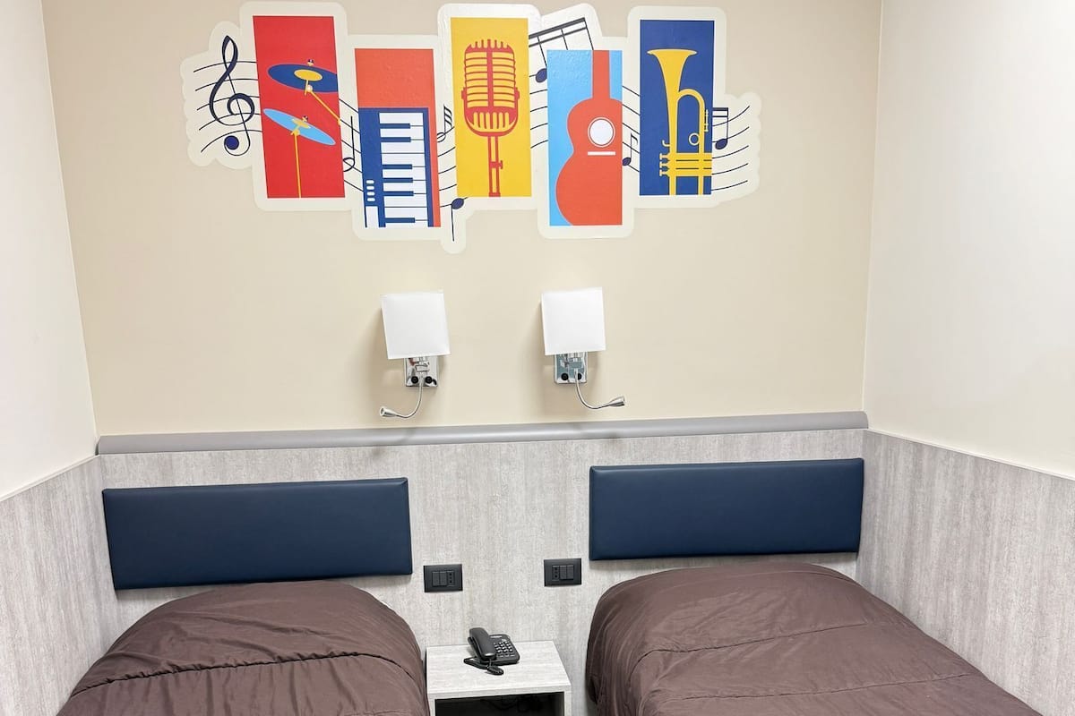 A hotel room with two beds, a wooden headboard, a small table with a phone, and wall art depicting musical instruments.