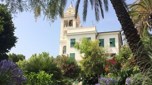 A white building with green shutters, surrounded by lush greenery and purple flowers. - Hotel Esperia (Sanremo)