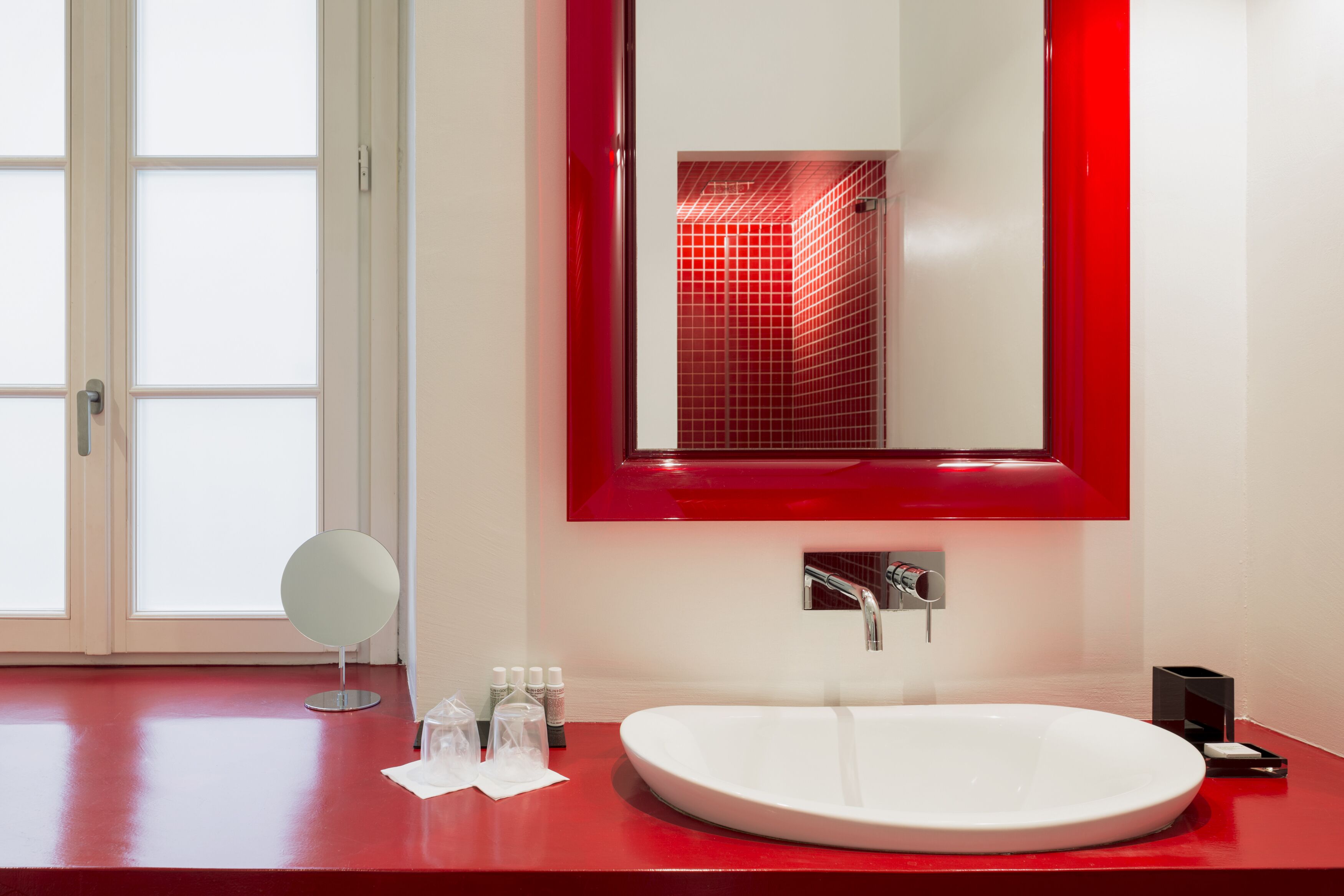 A modern bathroom with a white sink, a round mirror, and a red-framed mirror above the sink.