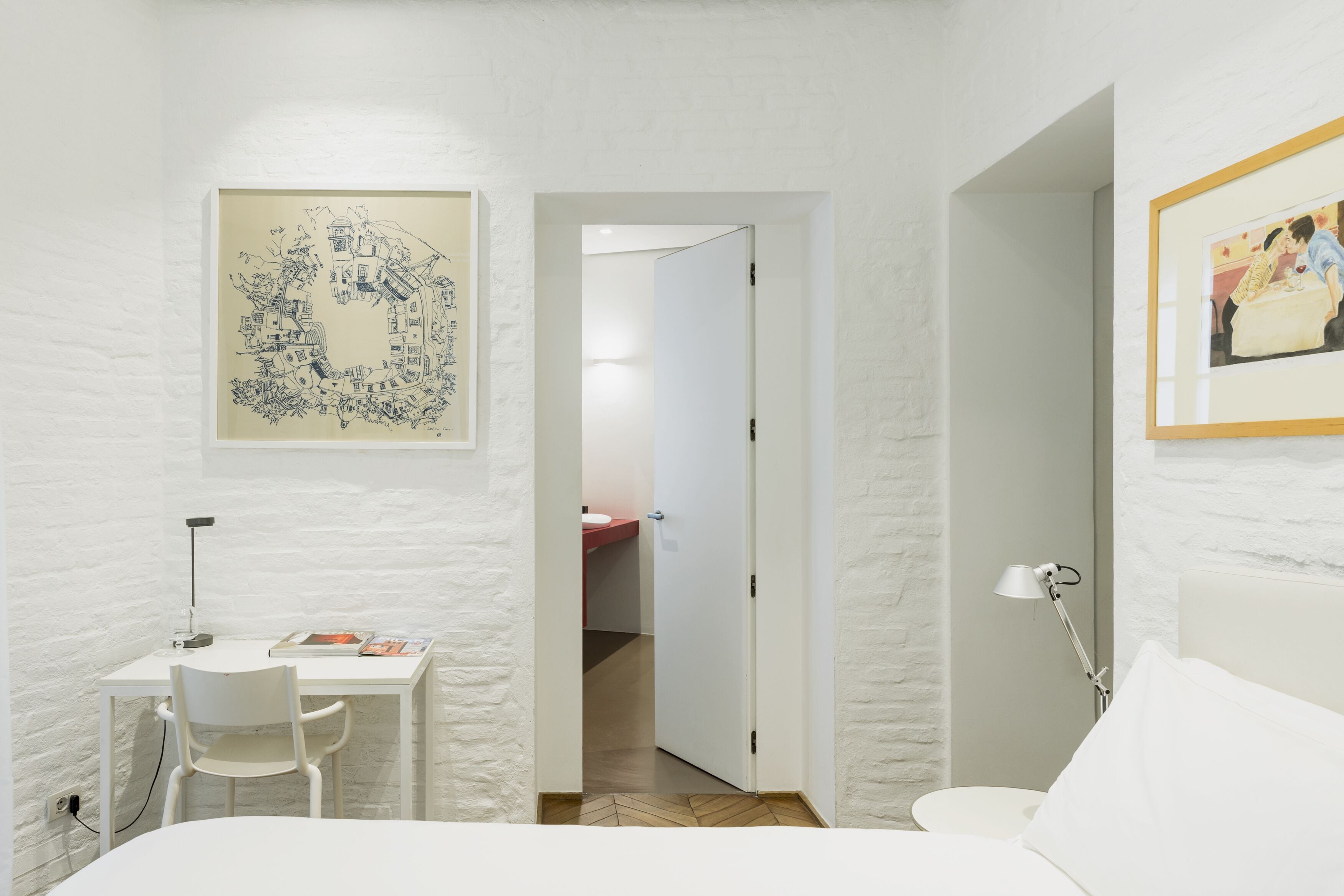 Una habitación con una cama, un escritorio con una silla, un cuadro en la pared y una puerta que da a otra habitación.
