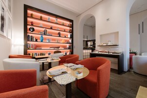 A modern interior with a central table, orange chairs, a bookshelf, and a kitchen area.