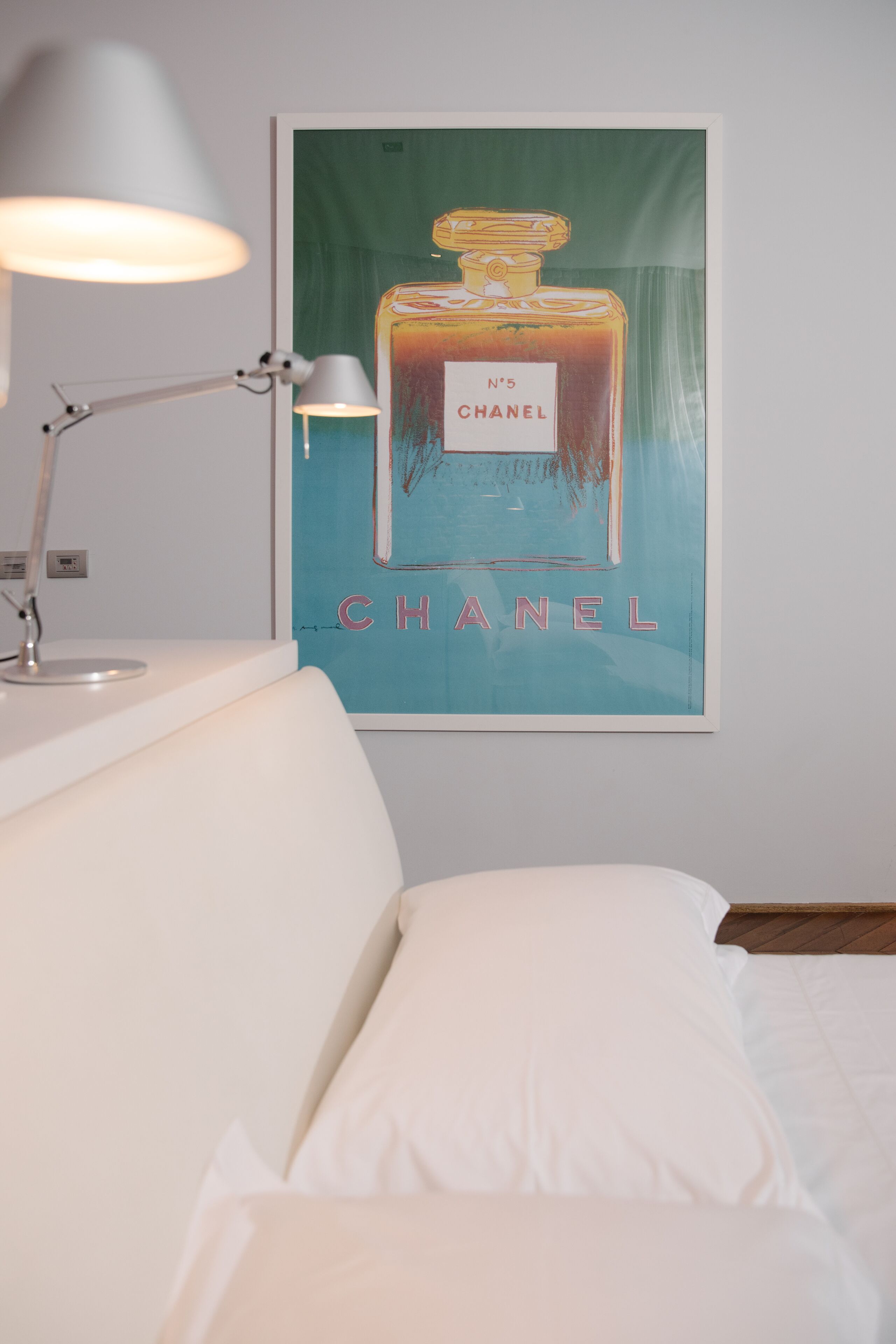 Quarto de hotel com uma cama, uma luminária branca e um cartaz de um frasco de perfume Chanel na parede.