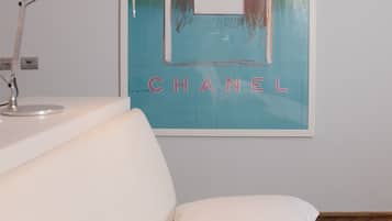 Quarto de hotel com uma cama, uma luminária branca e um cartaz de um frasco de perfume Chanel na parede.