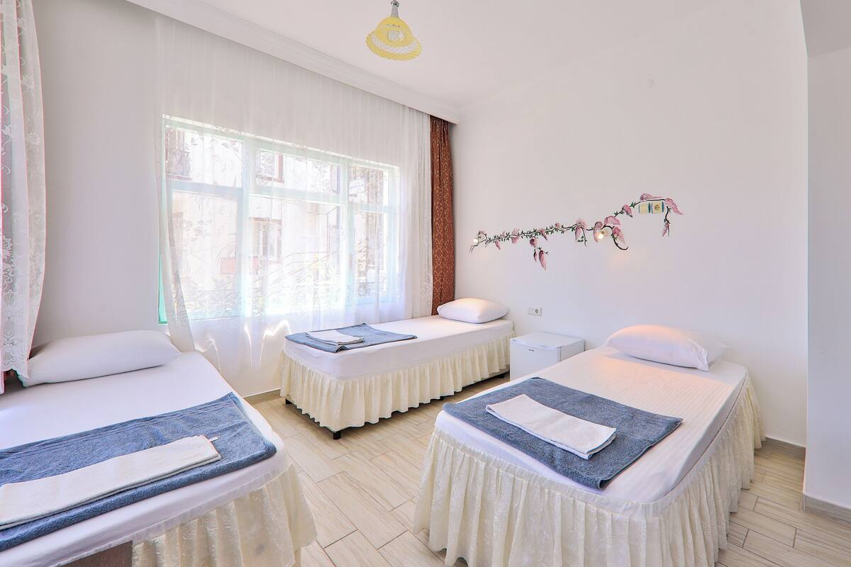 Una habitación de hotel con dos camas, una ventana con cortinas traslúcidas, un adorno en la pared y una mesita de noche pequeña.
