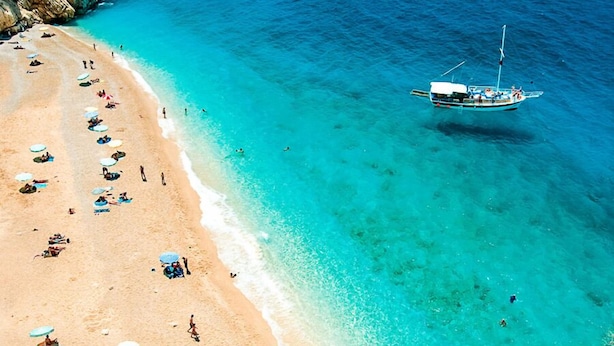 Uma praia com águas turquesa cristalinas, um barco ancorado perto da costa e pessoas tomando sol e nadando.