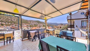 Um terraço com mesas e cadeiras, vista para uma vila no alto de uma colina, e um edifício com varanda.