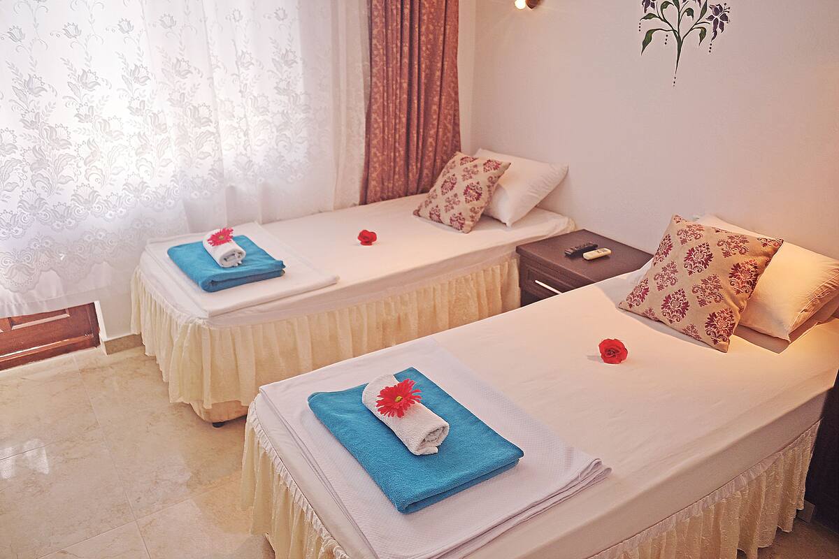 Dos camas individuales con sábanas blancas, toallas azules y almohadones decorativos. Cada cama tiene una mesita de noche con un control remoto.