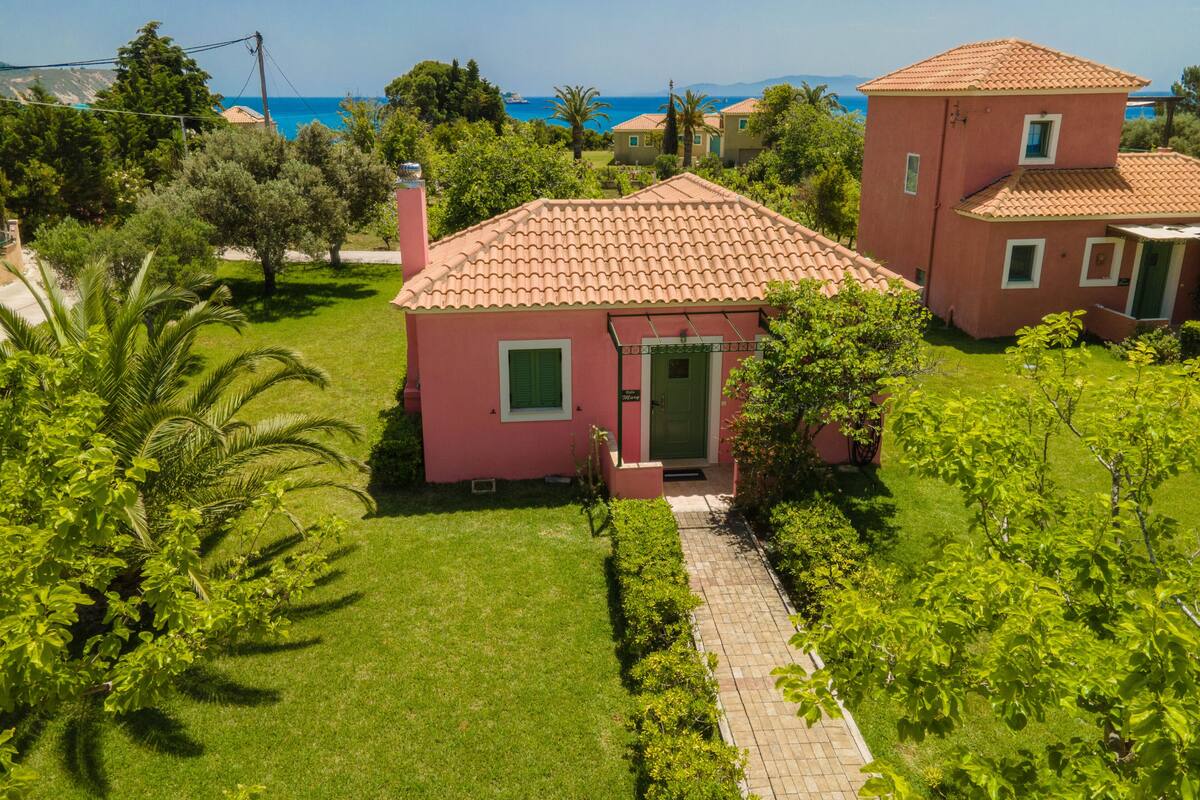 A pink villa with a tiled roof, surrounded by lush greenery and a clear view of the sea.