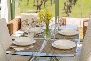 A dining table set with plates, glasses, and a vase of yellow flowers, with a view of trees and a bicycle in the background. - Monambeles Villas (Kefalonia)