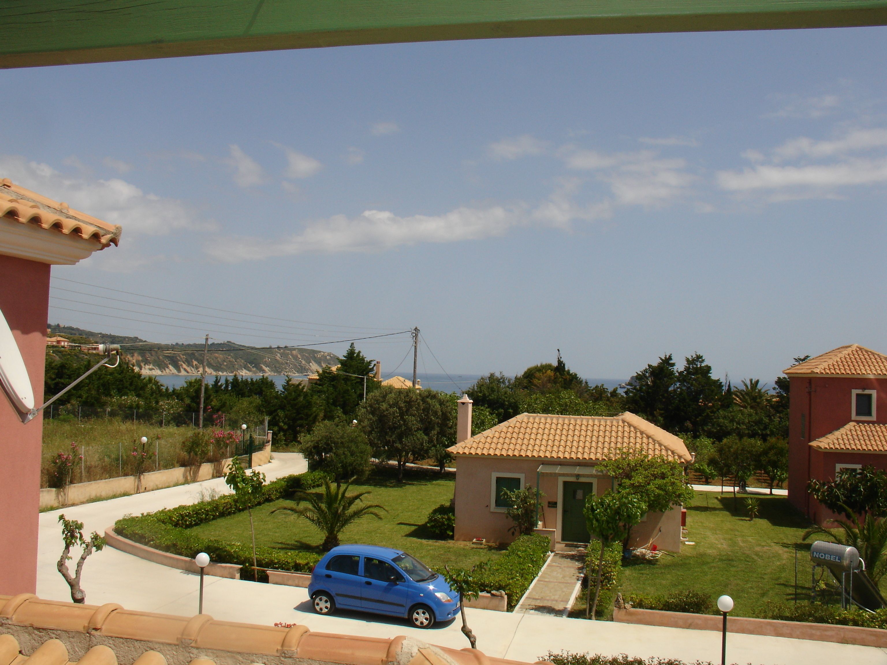 A residential area with a blue car parked in the driveway, houses with tiled roofs, and a clear view of the sea in the background.