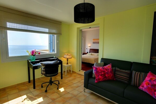 A room with a large window, a desk with a chair, a green sofa with colorful cushions, and a lamp on a small table. - Monambeles Villas (Kefalonia)