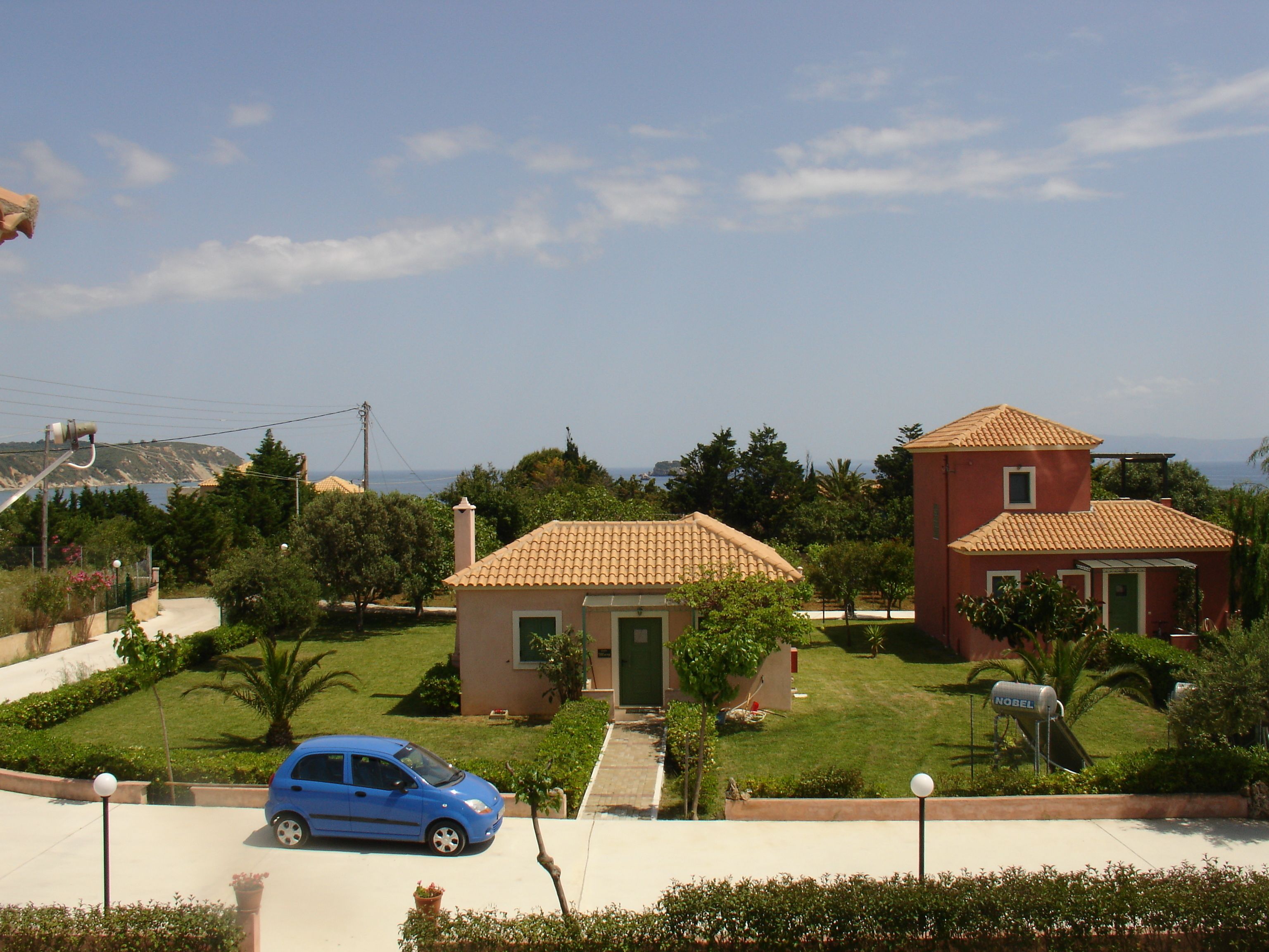 Zona residenziale con due case, un'auto blu parcheggiata davanti e una vista libera sul mare sullo sfondo.