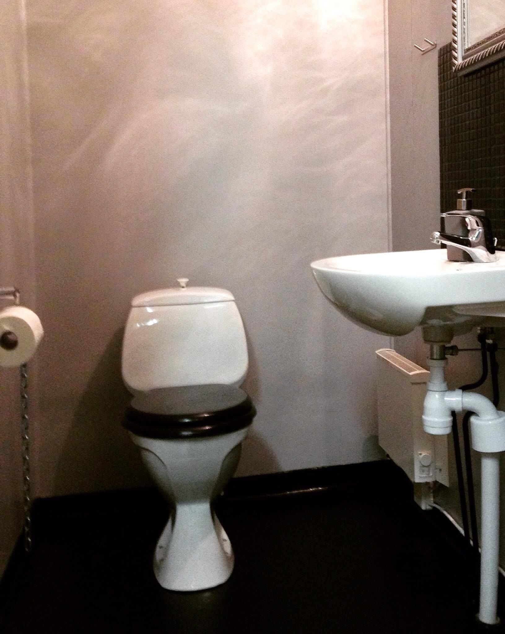 Un inodoro con asiento y tapa negros, un lavabo blanco con grifo y un dispensador de papel higiénico en la pared.