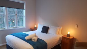 Una cama bien tendida con una colcha azul y toallas enrolladas, dos mesitas de noche con velas, una ventana con vistas a árboles y una silla en la esquina.