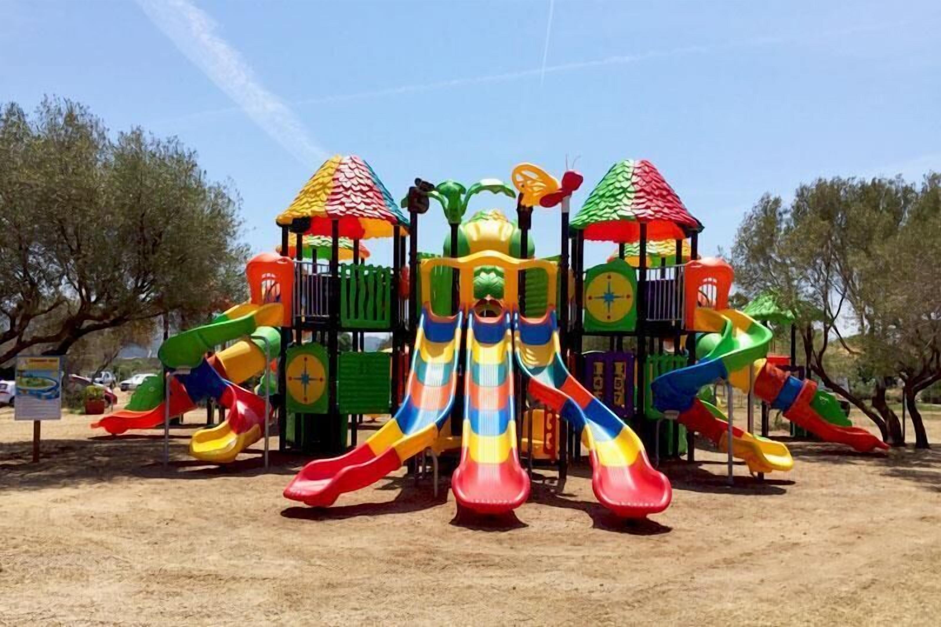 A playground with multiple slides and climbing structures, surrounded by trees.