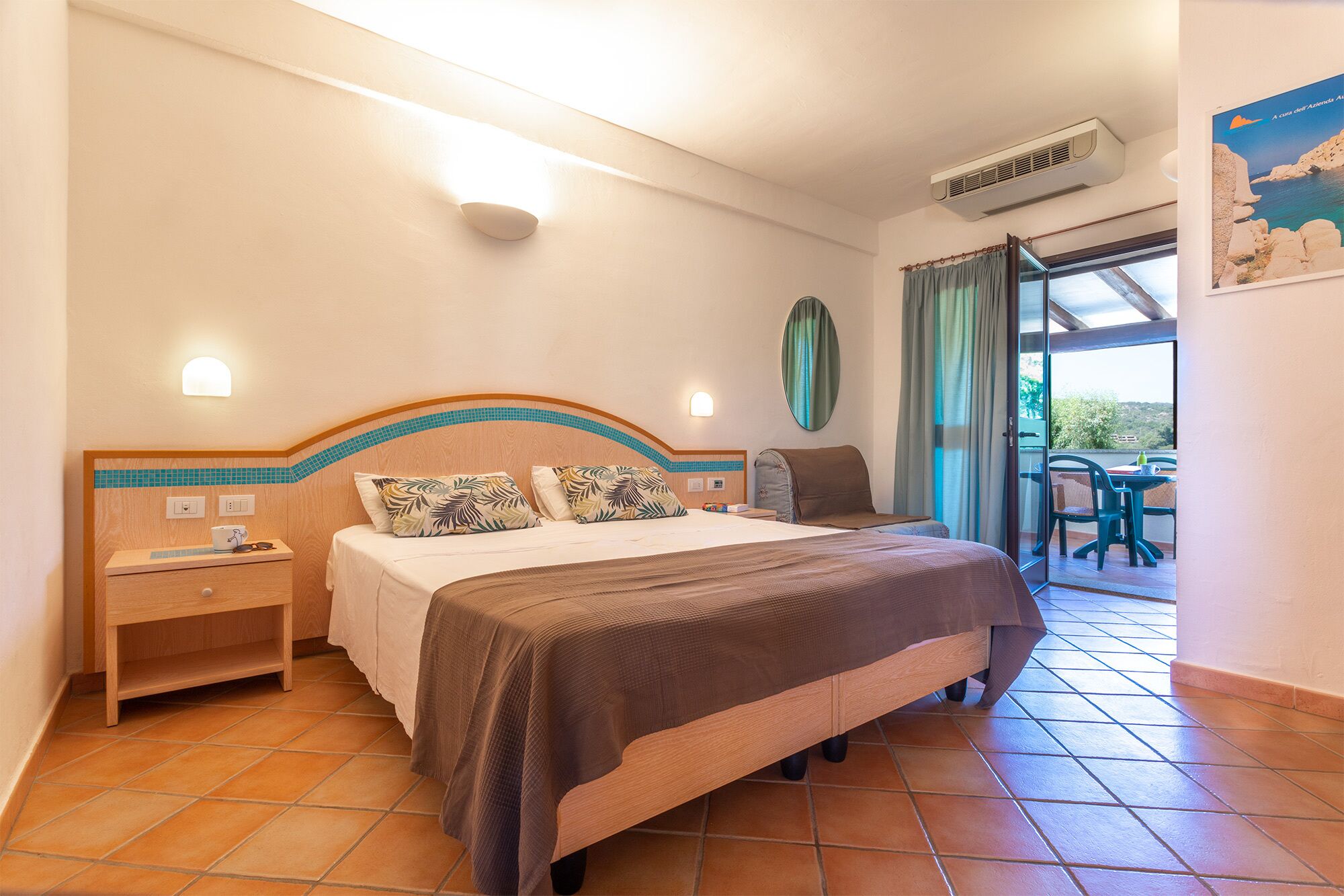 A hotel room with a bed, a nightstand, a table with chairs, a window with curtains, and an air conditioning unit.