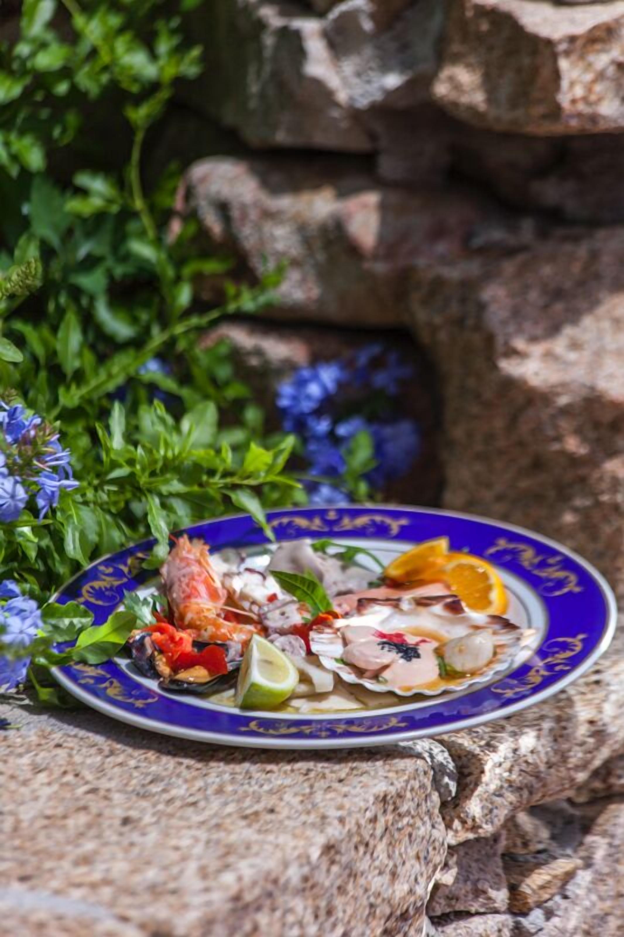 En tallerken med fisk og skaldyr på en blå og hvid mønstret tallerken, placeret på en stengulv med grønne planter og blomster i baggrunden.