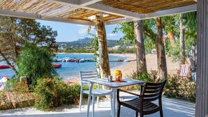Een overdekt terras met een tafel en stoelen, met uitzicht op een strand met boten en een bergachtige achtergrond.